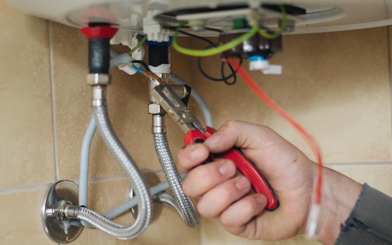 Hand using pliers on the fittings and braided hoses under a water heater